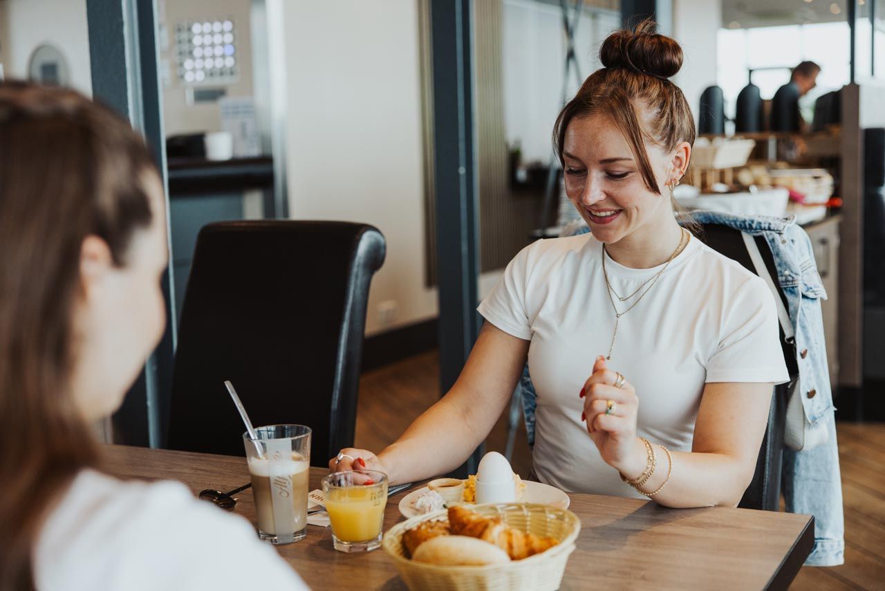 Zwei Frauen unterhalten sich bei Kaffee und Gebäck an einem Café-Tisch; im Hintergrund ist ein Laptop zu sehen.