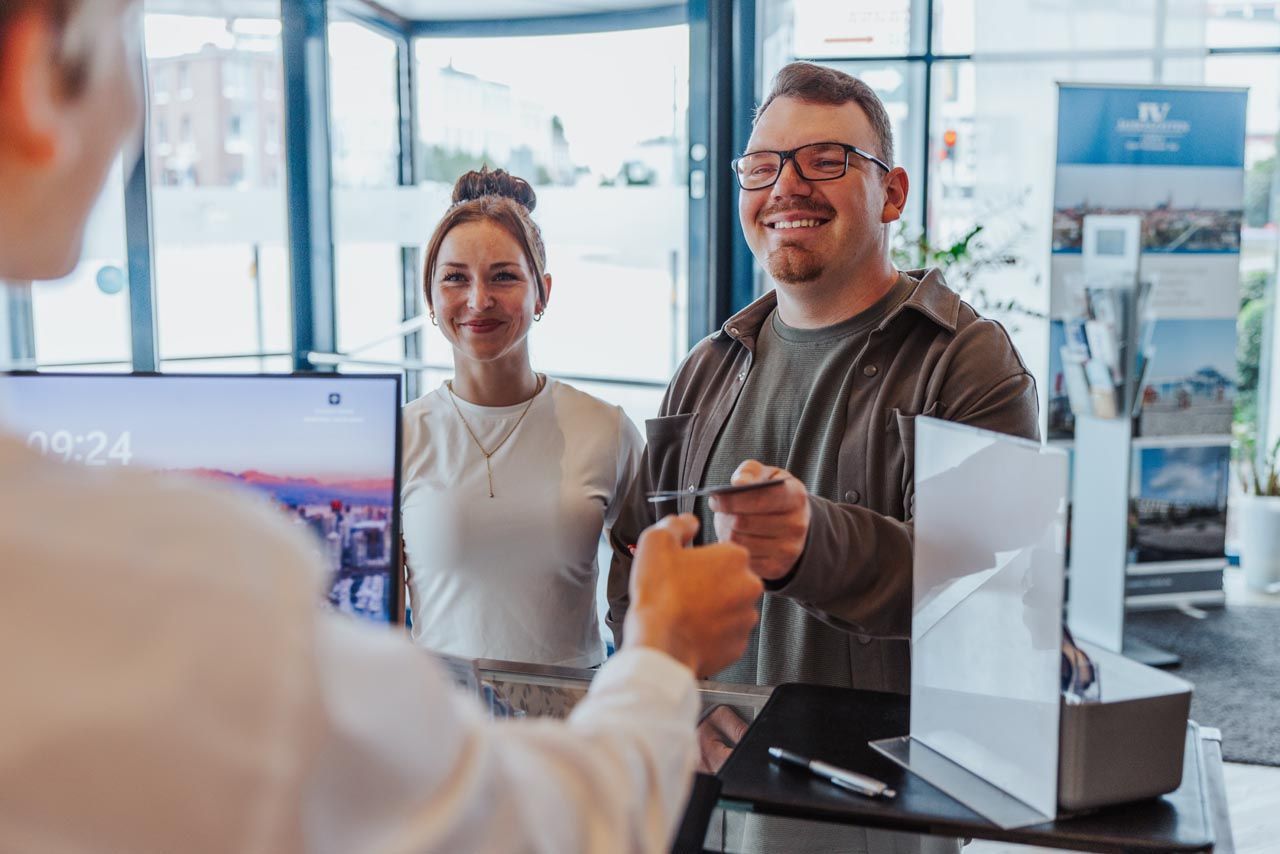 Zwei lächelnde Personen an einem Empfangstresen erhalten in einer hellen Bürolobby eine Karte von Mitarbeitern.