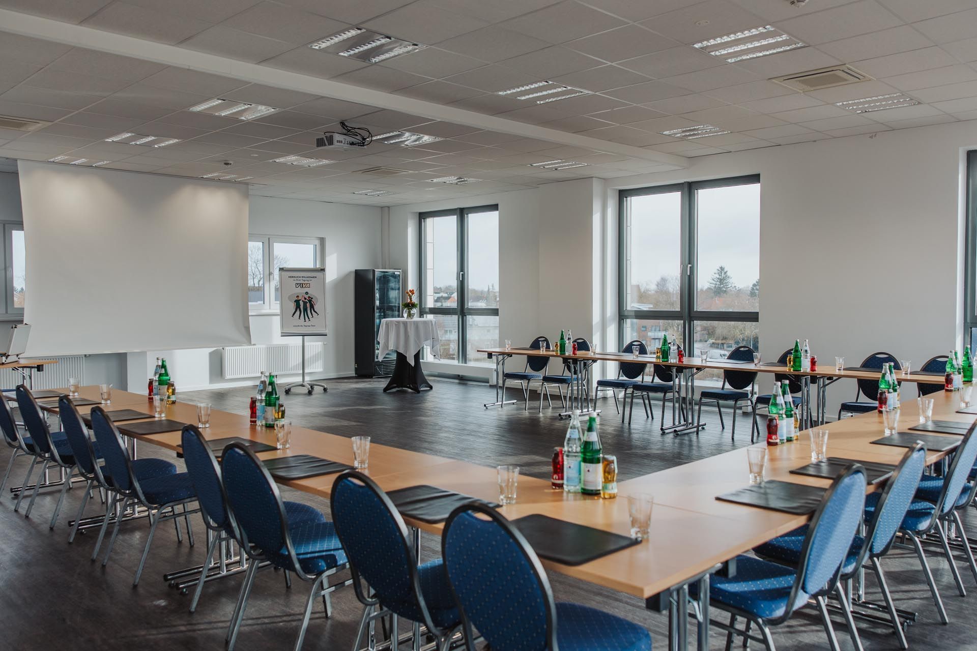 Leerer Konferenzraum mit U-förmigen Tischen, blauen Stühlen und Flaschengetränken an großen Fenstern