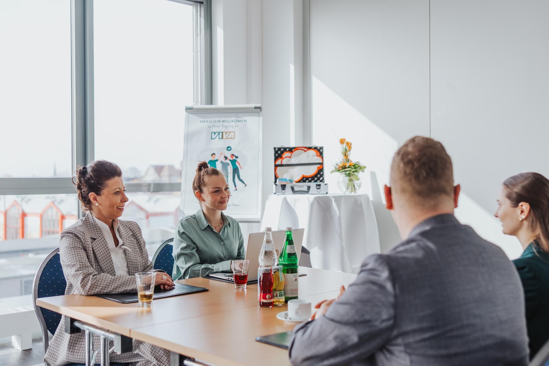 Vier Personen sitzen in einem hellen Büro um einen Tisch mit Getränken und Papieren.