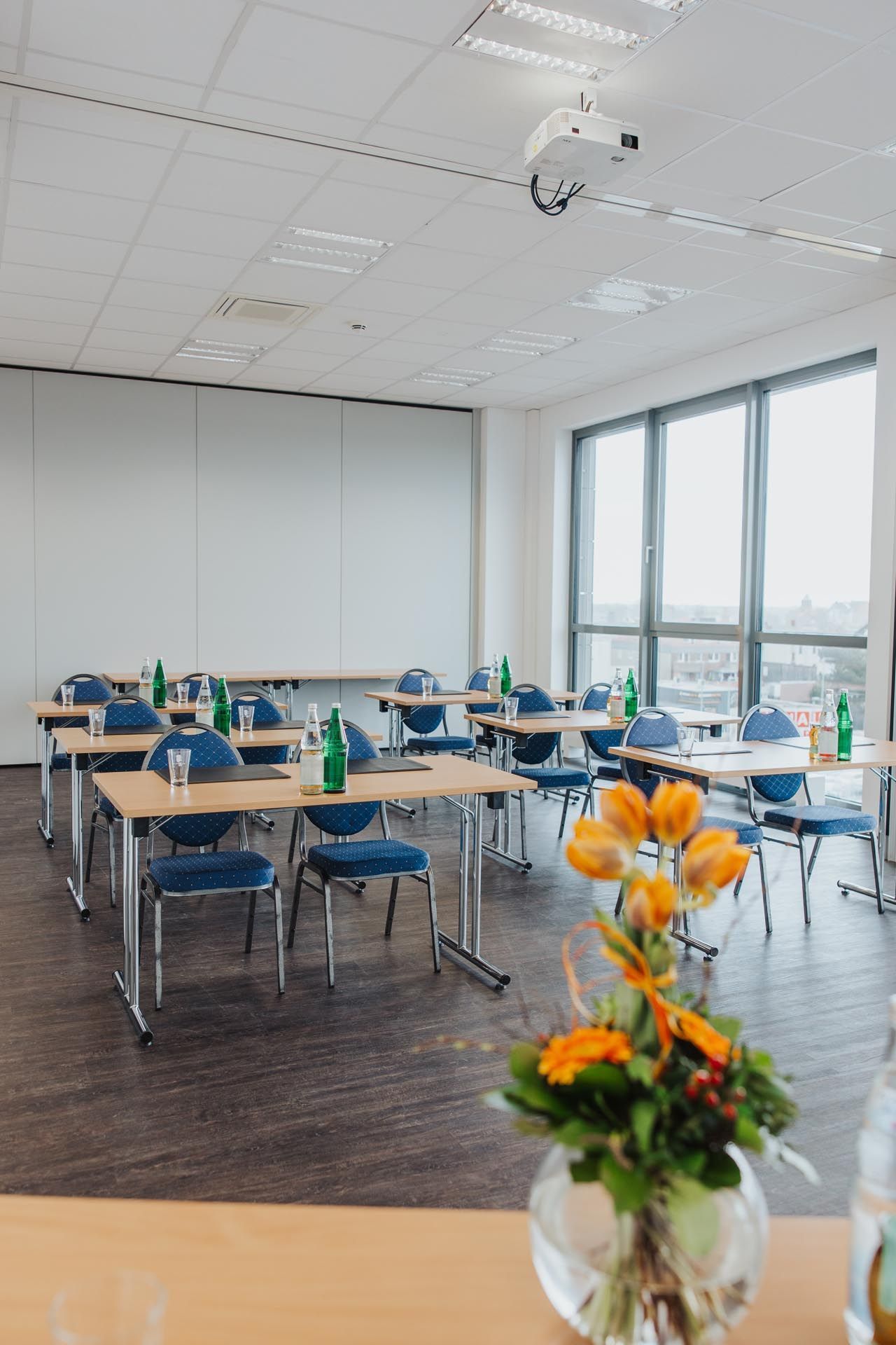 Leeres Klassenzimmer mit Tischen und blauen Stühlen, großen Fenstern und einer Vase mit orangefarbenen Blumen im Vordergrund