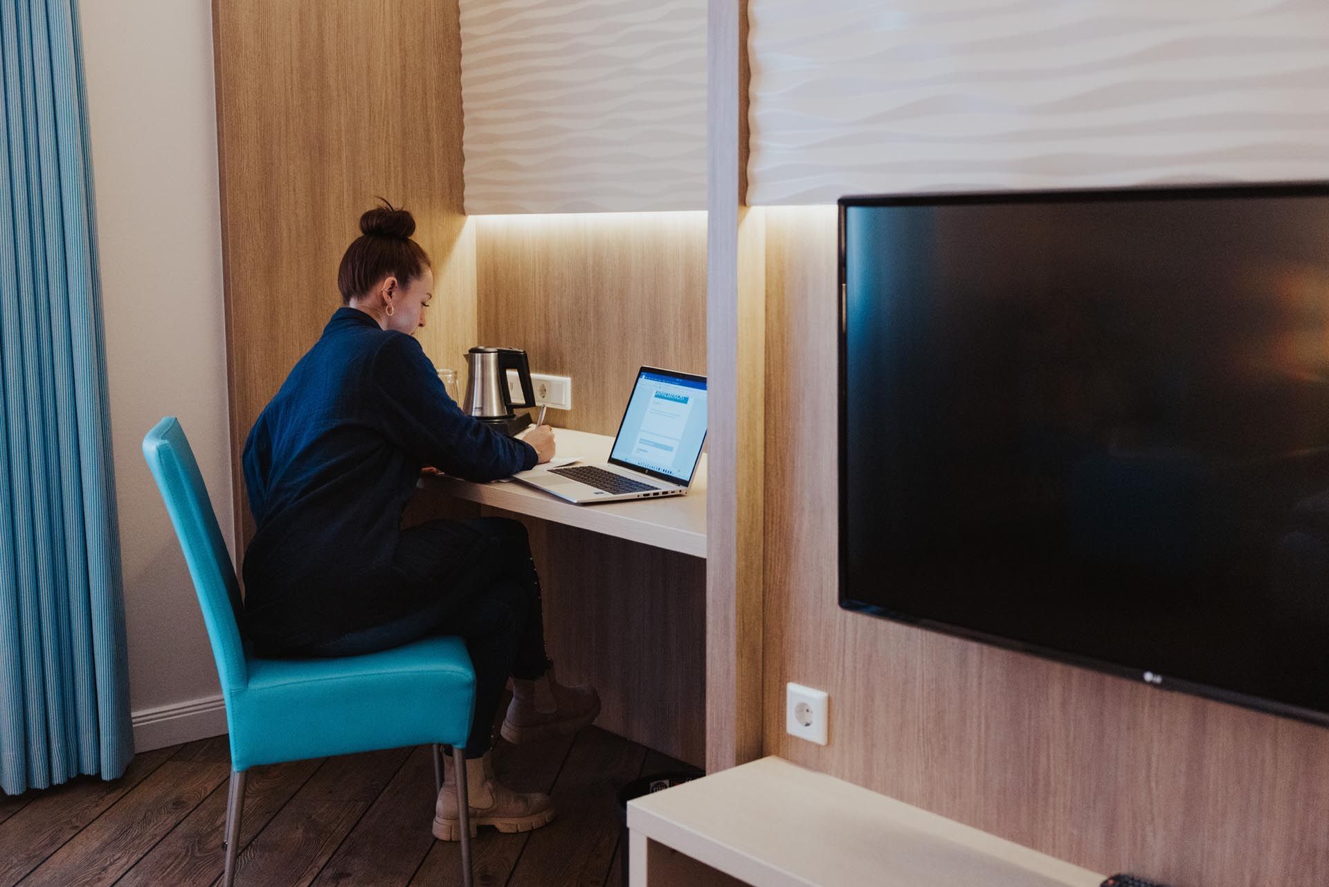Eine Frau arbeitet an einem Laptop an einem Schreibtisch in einem Hotelzimmer mit Fernseher und einem türkisfarbenen Stuhl.