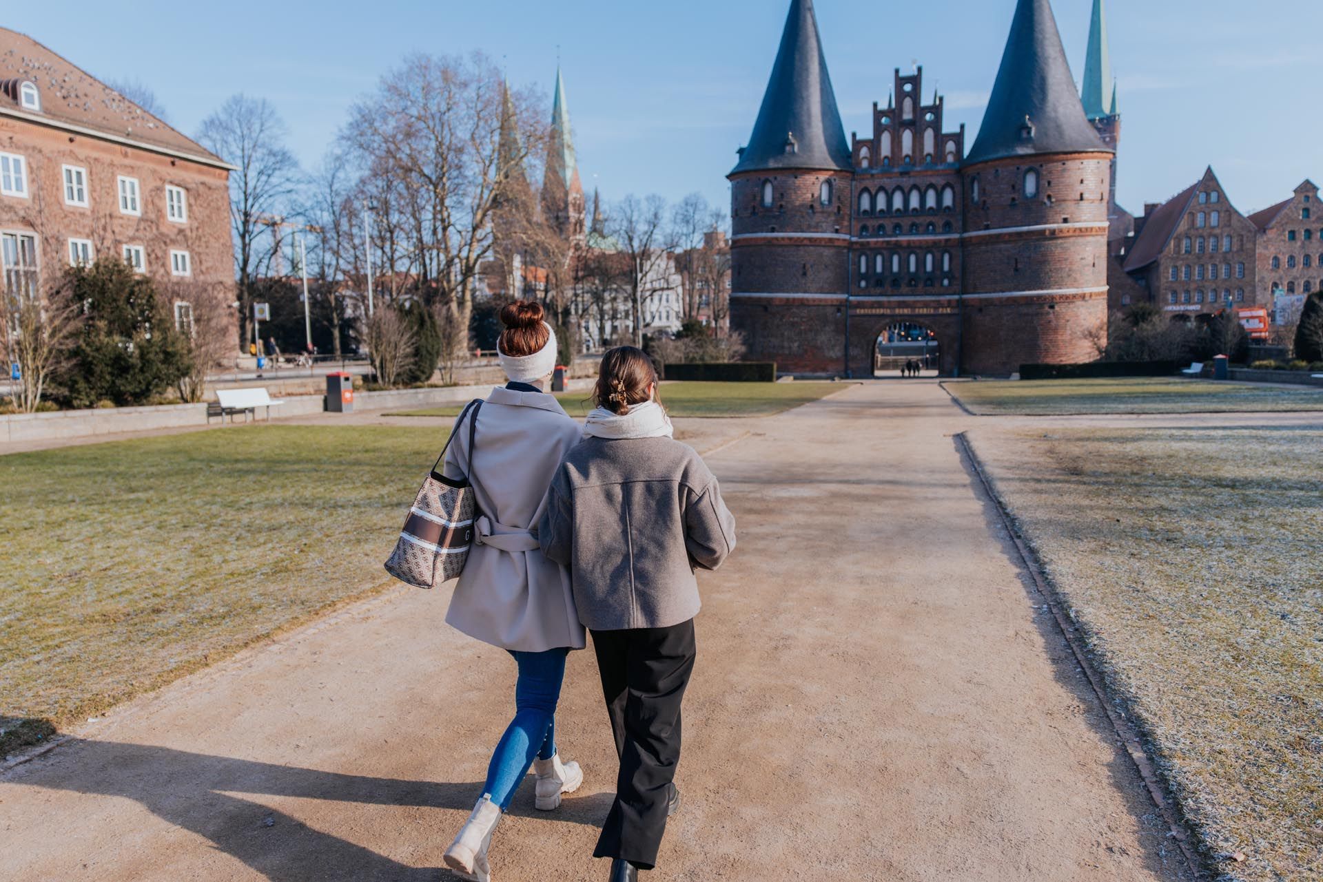 Zwei Personen gehen an einem sonnigen Tag auf ein Backsteintor mit Bogentürmen zu.