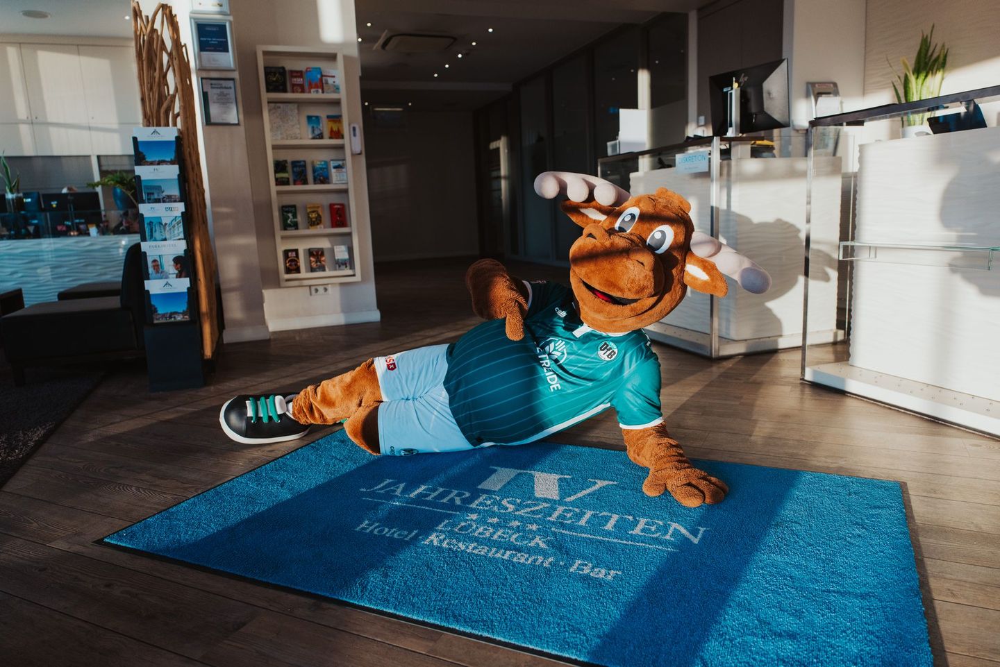 Ein orangefarbenes Maskottchen liegt in einer hellen Lobby auf einer blauen Matte;