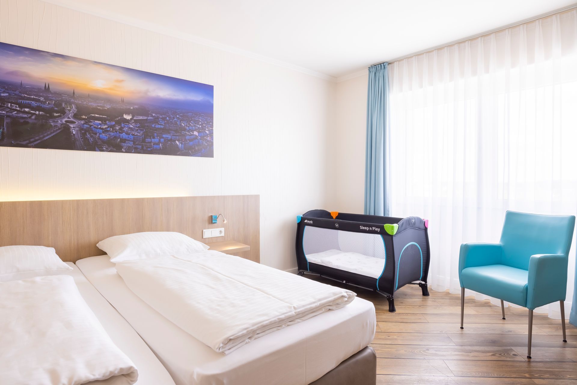Hotelzimmer mit zwei Betten, Kinderbett, blauem Stuhl und Stadtbild.