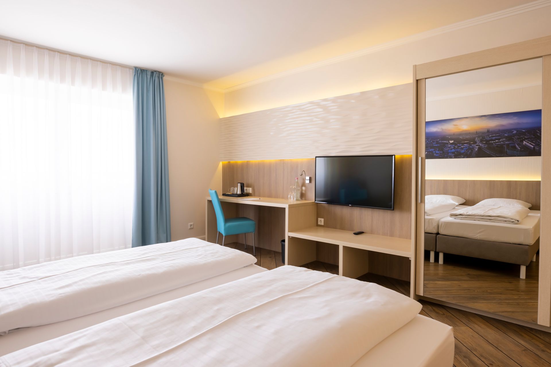 Hotelzimmer mit zwei Betten, Schreibtisch, Fernseher und großem Spiegel, in dem sich die Betten und die Kunstwerke spiegeln.