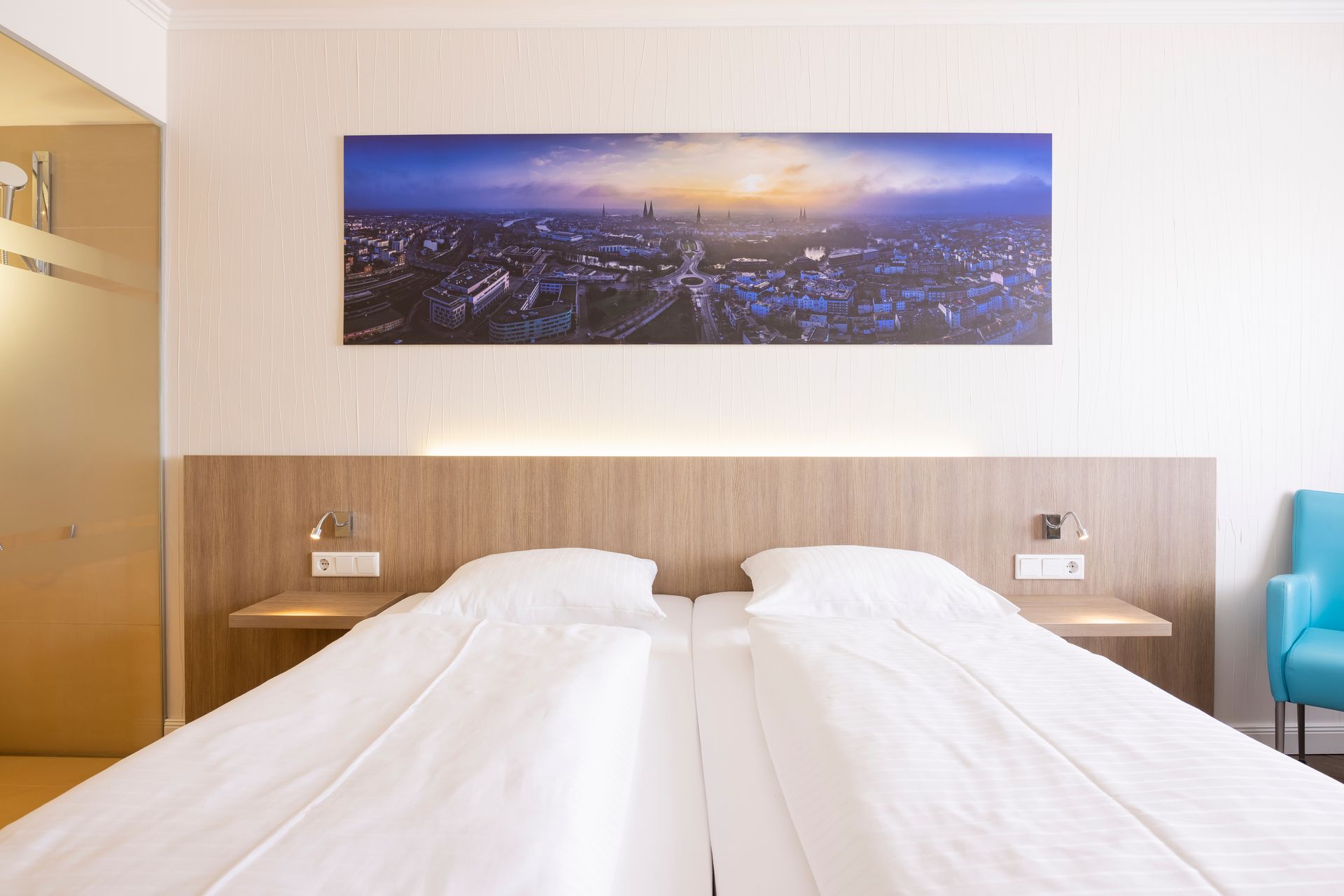 Hotelzimmer mit zwei Einzelbetten, Kopfteil, Nachttischen, Stadtbild und einem blauen Stuhl.