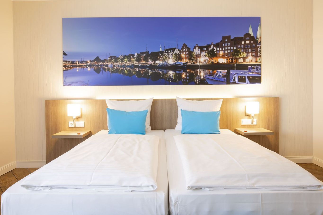 Helles Hotelzimmer mit zwei Einzelbetten, blauen Kissen und einem Stadtpanoramafoto über dem Kopfteil.