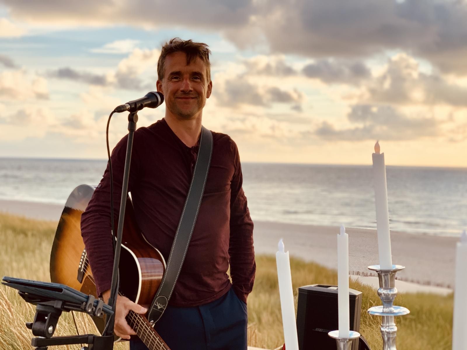 Mann mit Gitarre spielt bei Sonnenuntergang am Strand, umgeben von Kerzen und Tontechnik; er lächelt in die Kamera.