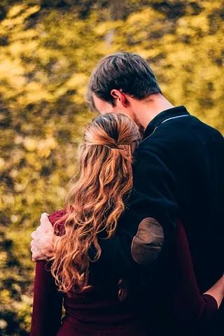 Pareja abrazándose al aire libre, el cabello rizado de la mujer, el brazo del hombre rodeándola. Colores otoñales, fondo desenfocado.