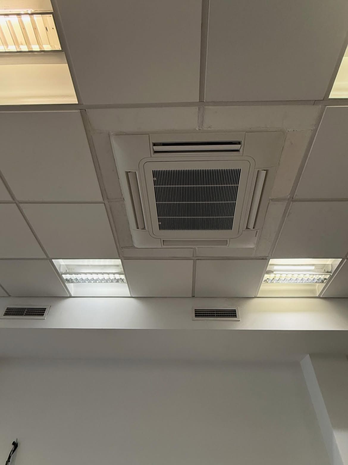 Techo de oficina con paneles de luz empotrados y una rejilla de ventilación central sobre una pared blanca.