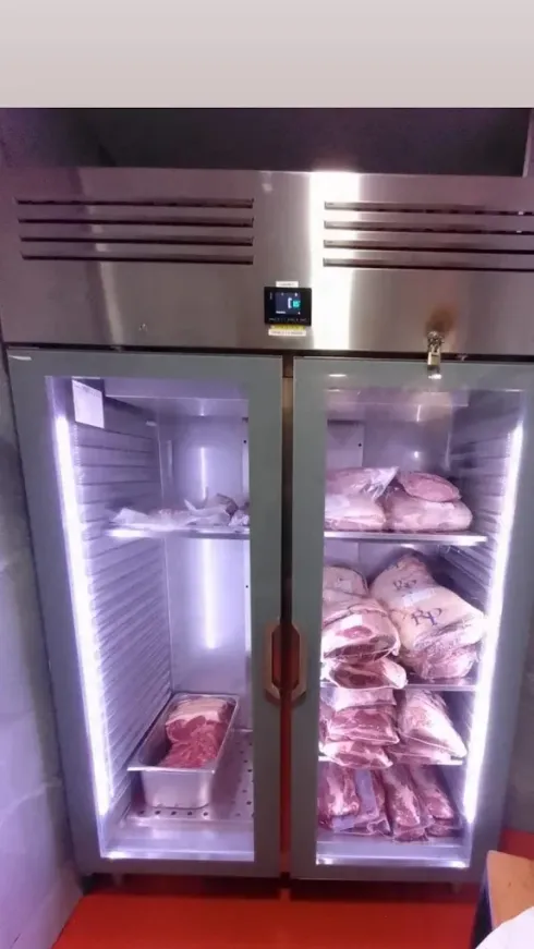 Refrigerador de acero inoxidable con puertas de vidrio que muestran bandejas de carne cruda en su interior