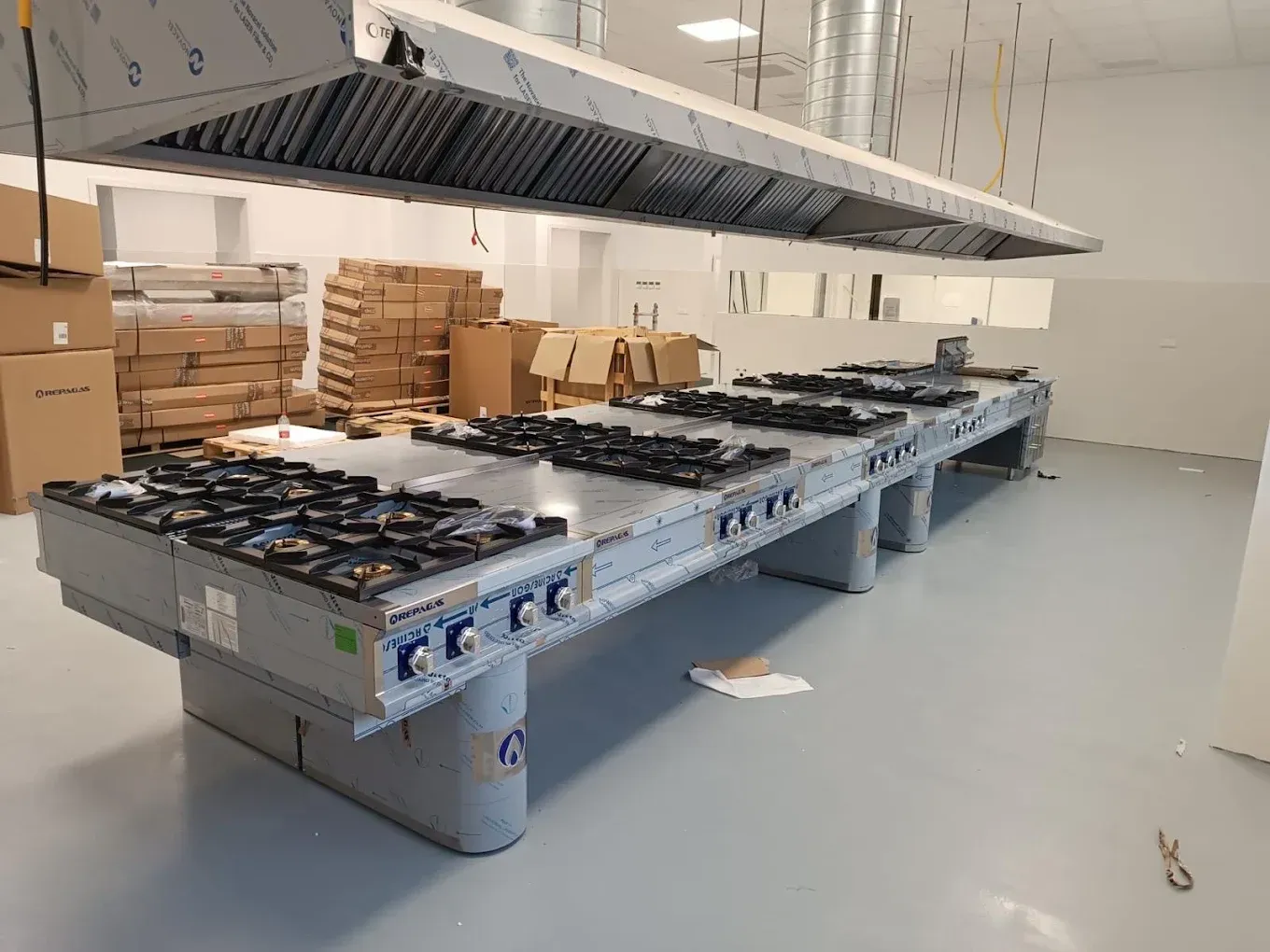 Cocina industrial con una larga línea de cocción, campanas extractoras en el techo, cajas apiladas y un suelo gris.