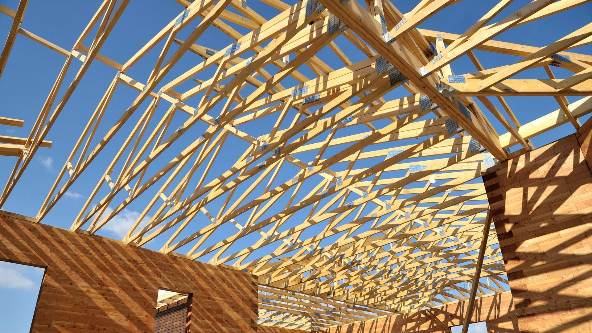 Charpente de toit en bois se détachant sur un ciel bleu, encadrant l'intérieur d'un bâtiment en construction.