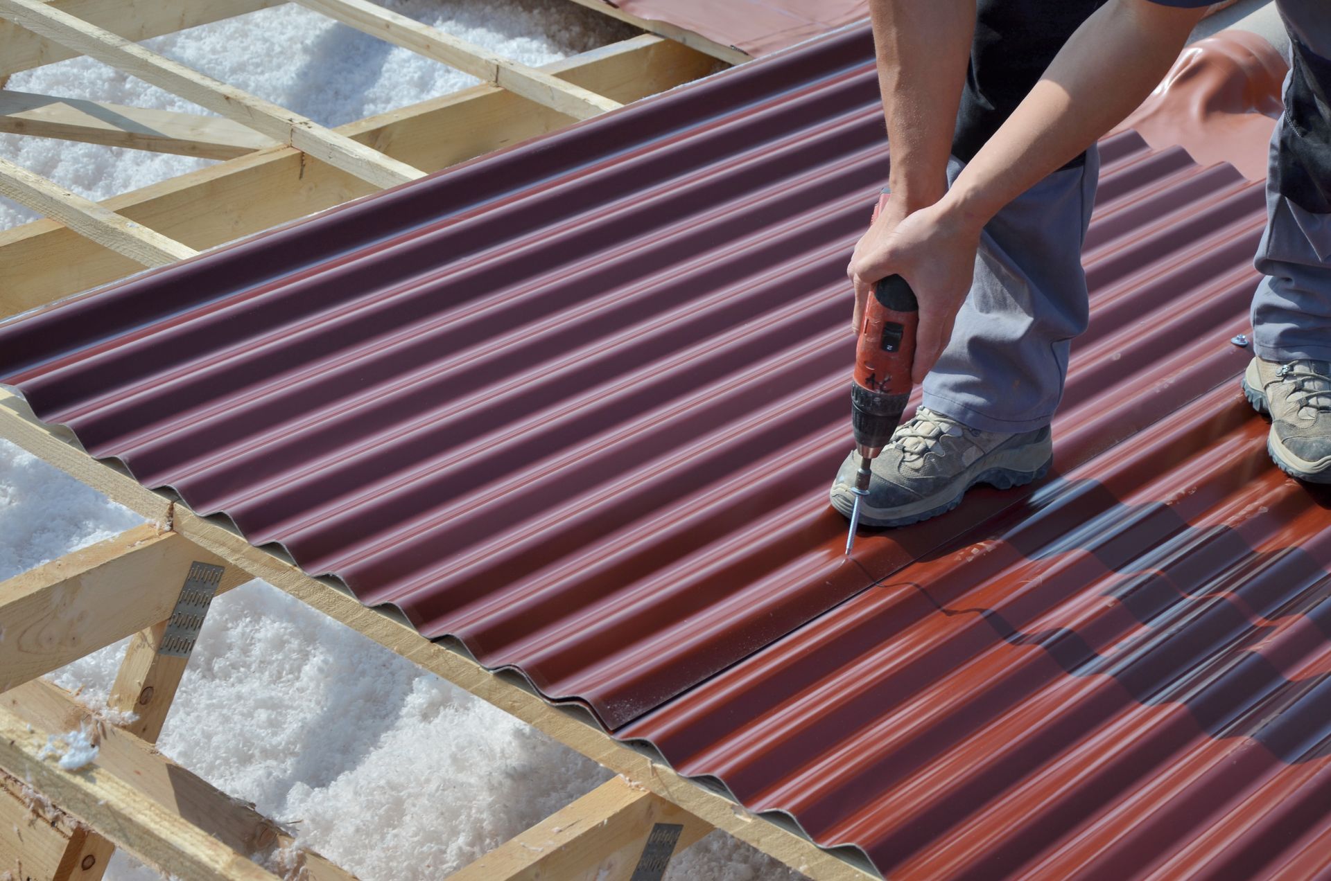 Une personne utilise une perceuse pour fixer des tôles de toiture ondulées à des chevrons en bois.
