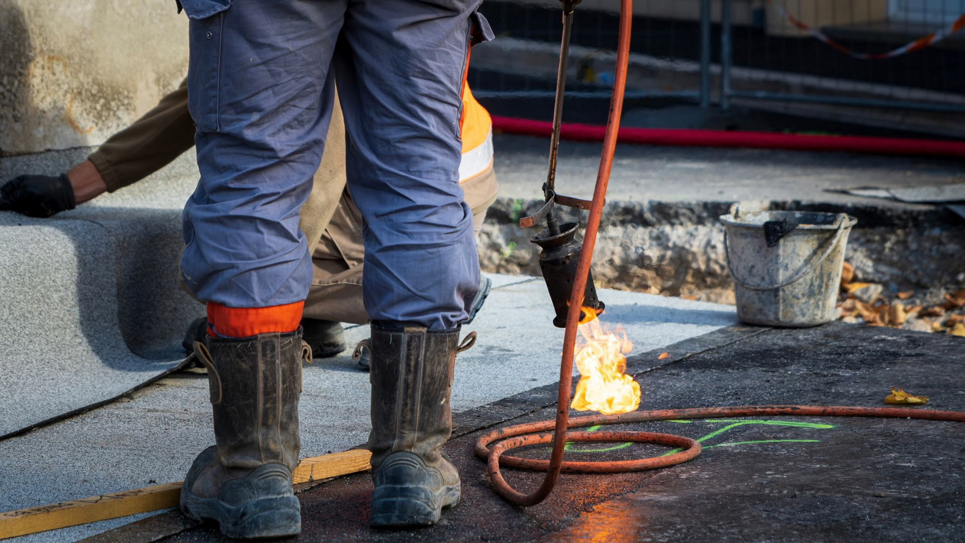 Un ouvrier du bâtiment utilise une torche pour travailler sur un toit, portant des bottes et un équipement de sécurité.