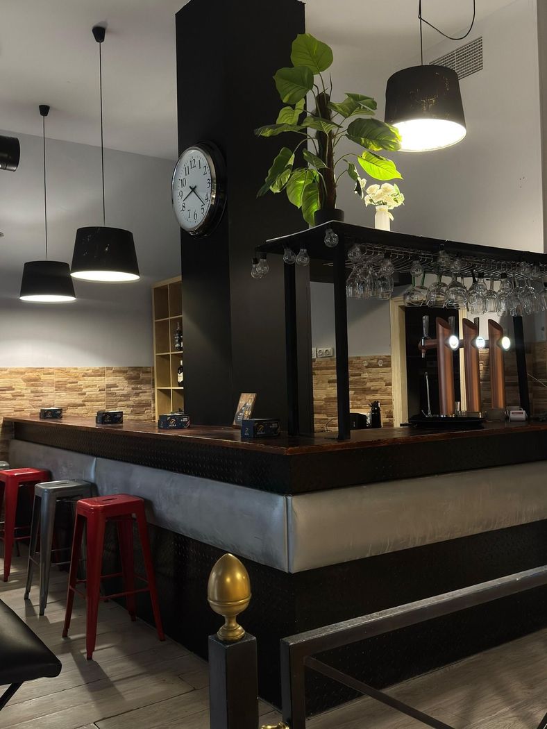 Interior de bar moderno con lámparas colgantes, taburetes rojos y una barra central con vasos y plantas colgantes.