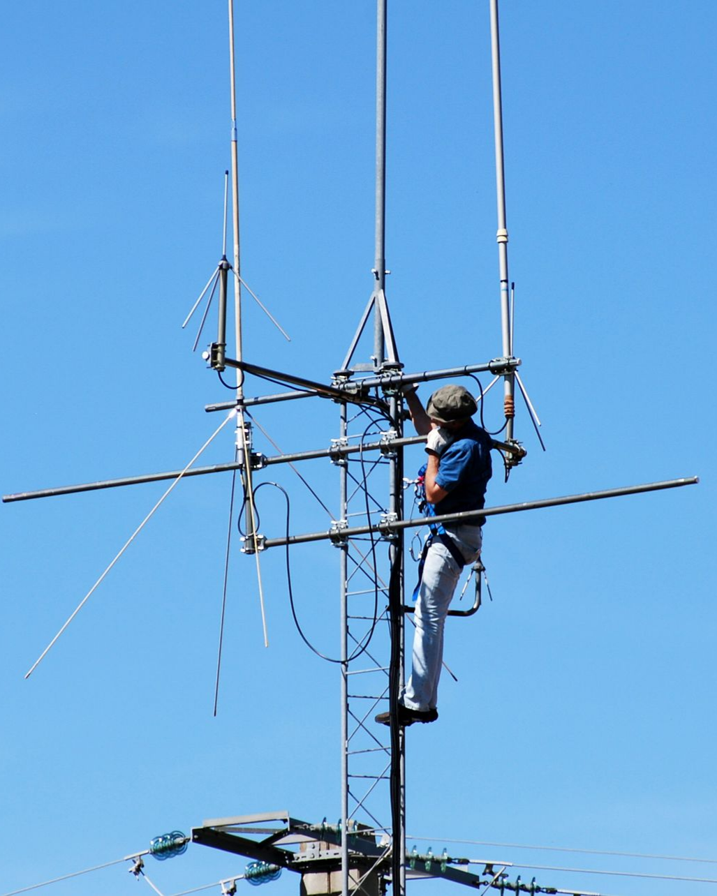 Un homme, perché sur une tour radio, ajuste les antennes sur fond de ciel bleu.
