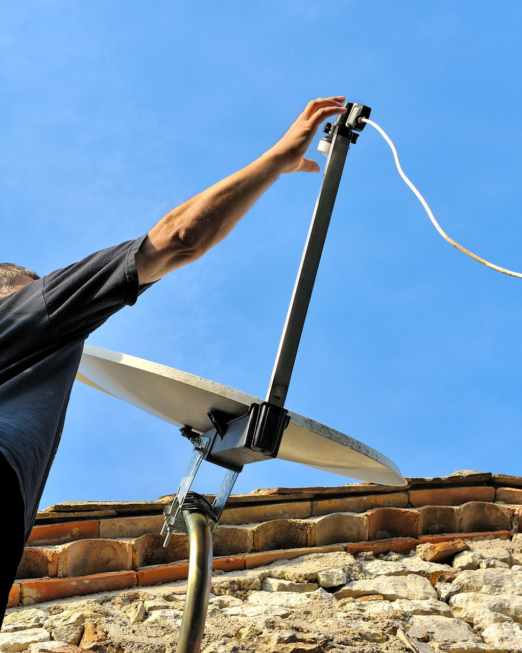 Une personne ajuste une antenne parabolique sur un toit de briques, sur fond de ciel bleu.