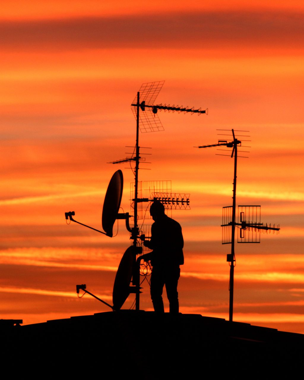 Silhouette d'une personne ajustant des antennes sur un toit, se détachant sur un coucher de soleil.