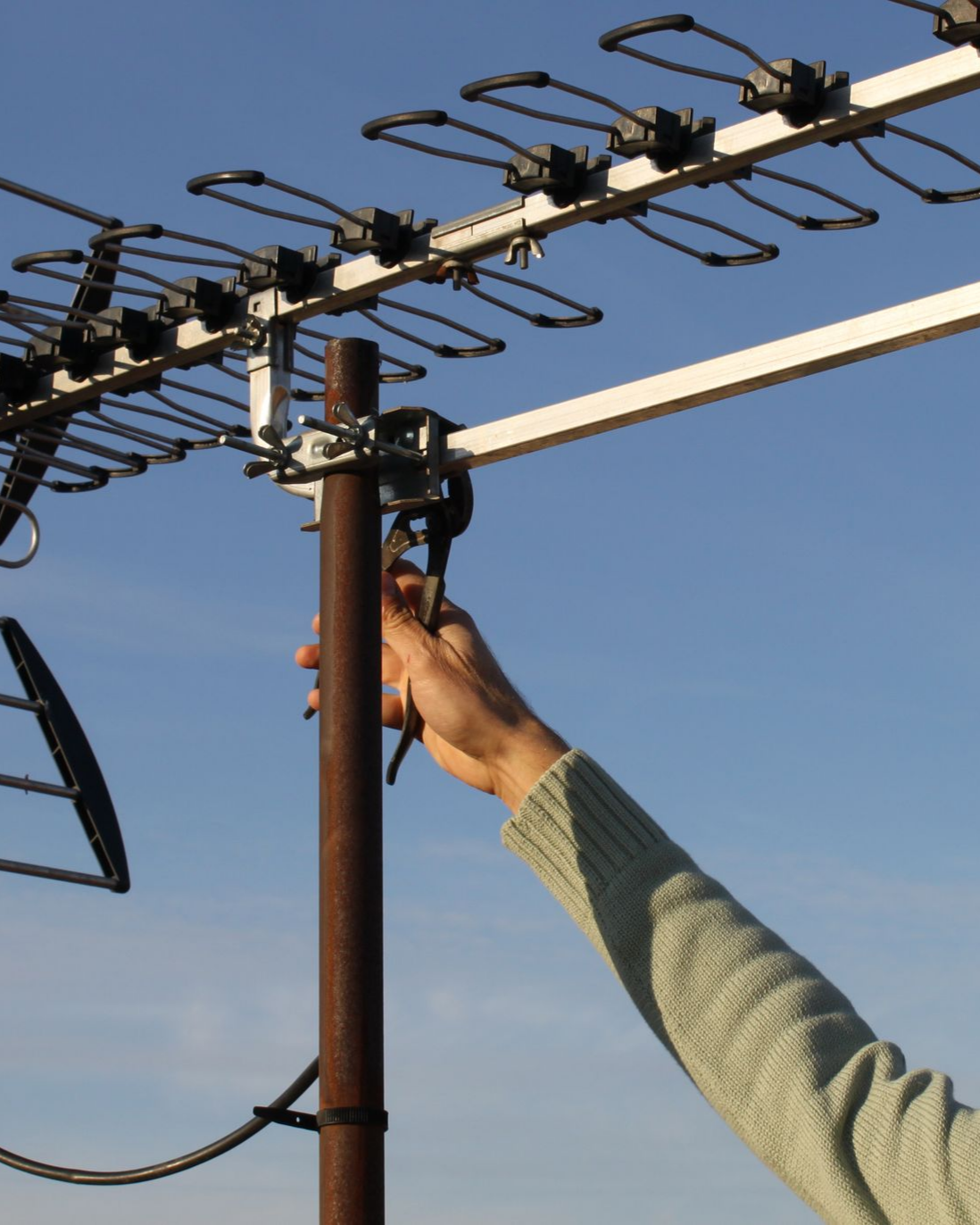 Une main serre un collier de serrage sur une antenne de toit. Ciel bleu en arrière-plan.