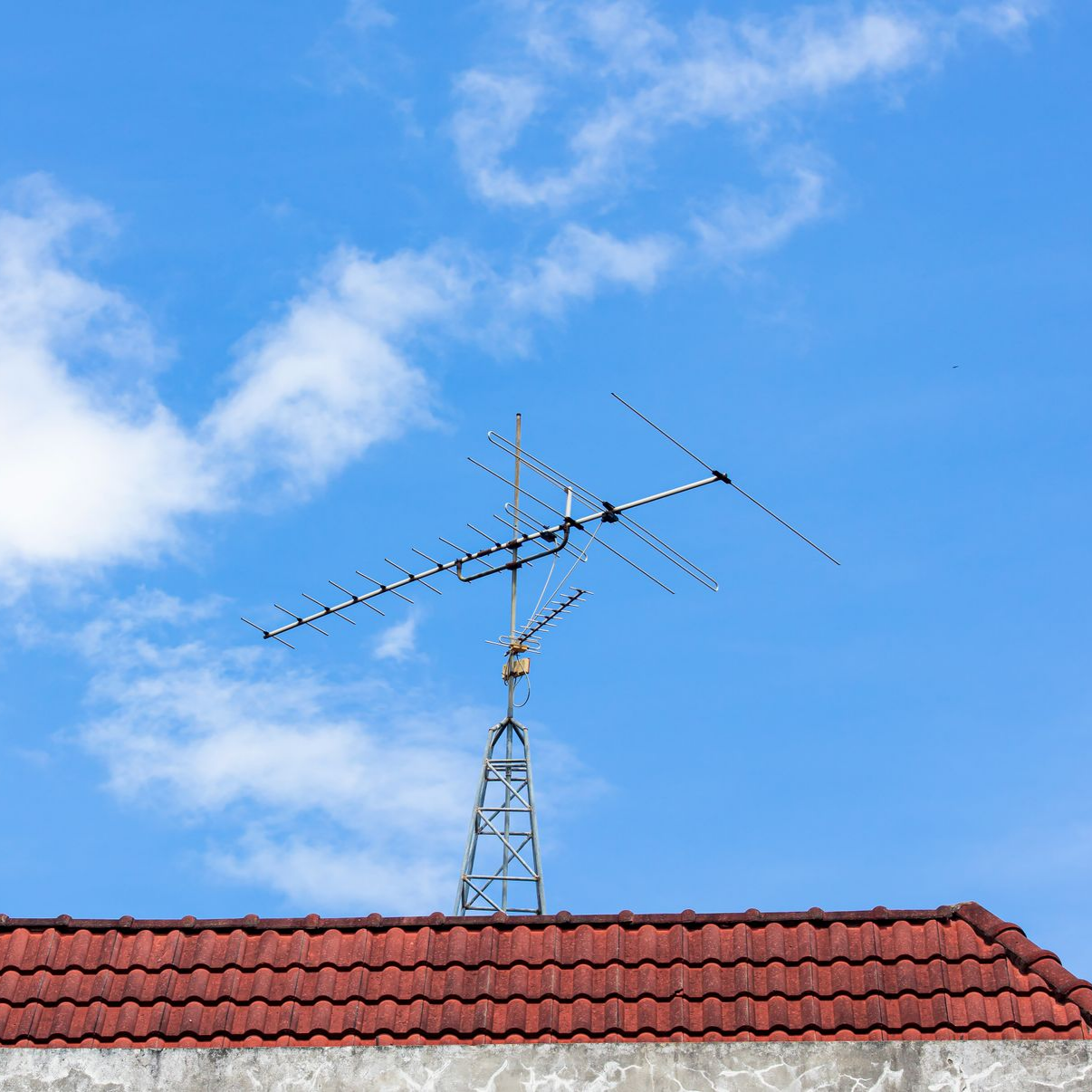 Antenne de télévision sur un toit de tuiles rouges, se détachant sur un ciel bleu parsemé de nuages ​​blancs.
