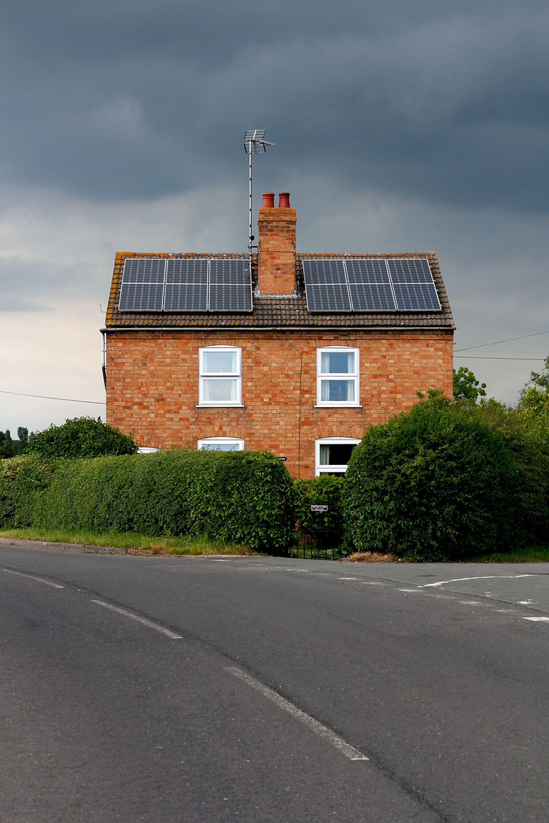 Maison en briques avec panneaux solaires sur le toit, haies, cheminée et antenne par temps gris.