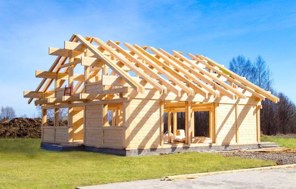 Auf einem grasbewachsenen Grundstück unter klarem, blauem Himmel steht ein teilweise errichtetes Holzhausgerüst mit Dachsparren.