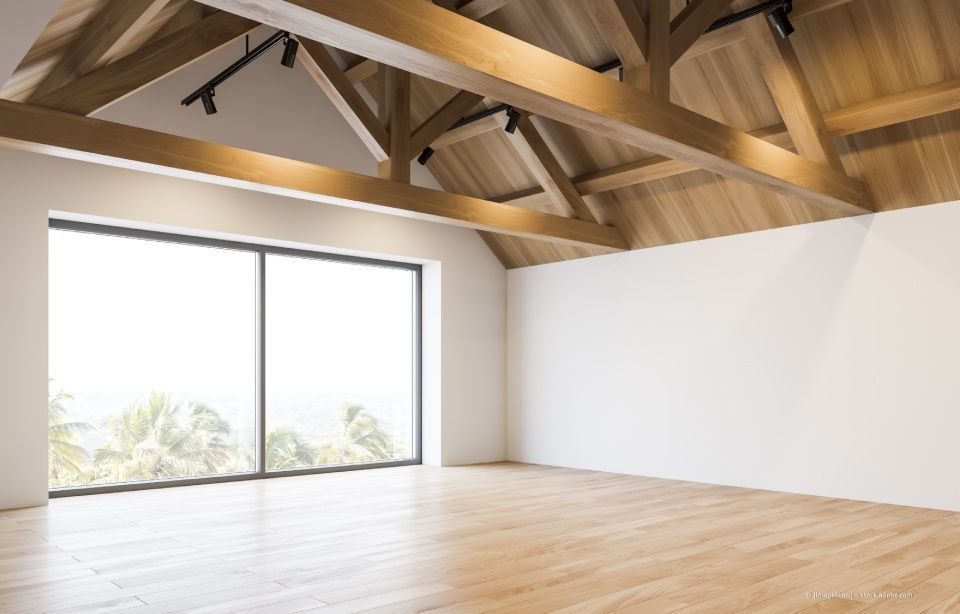 Ein leerer Raum mit weißen Wänden, hellem Holzboden und einer gewölbten Decke mit freiliegenden Holzbalken und Schienenstrahlern.