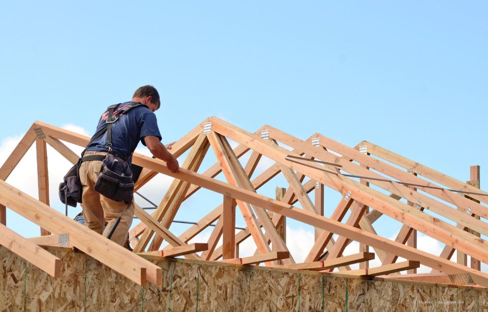 Ein Arbeiter mit Werkzeuggürtel justiert vor strahlend blauem Himmel einen Holzfachwerkträger am Dachstuhl eines Hauses.
