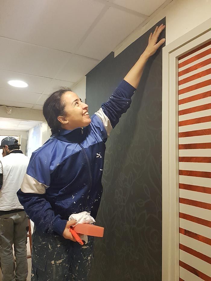 Mujer con chaqueta azul que se acerca a la pared, sosteniendo herramientas, y aplica pintura oscura en una habitación.