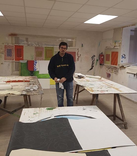 Hombre en un estudio de arte junto a una gran obra de arte. Mesas con materiales de arte y coloridas pinturas abstractas en las paredes.