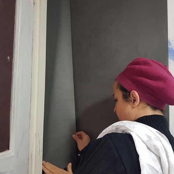 Mujer con sombrero rojo aplicando papel tapiz gris oscuro en una pared al lado de una puerta blanca.