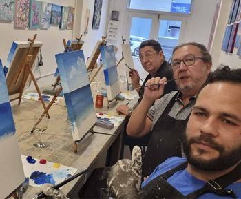 Personas pintando en caballetes en una clase de arte. Están trabajando en pinturas de cielo azul.