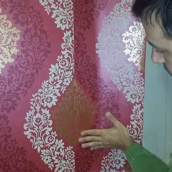 Un hombre inspecciona un papel tapiz floral rojo y dorado dañado en una pared.