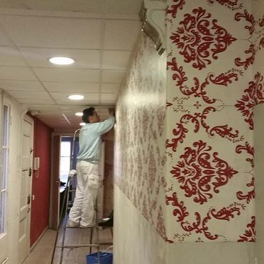 Una persona en una escalera instala un papel tapiz con un patrón rojo y blanco en un pasillo.