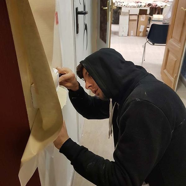 Hombre con una sudadera con capucha negra instalando papel tapiz en una pared dentro de un edificio.
