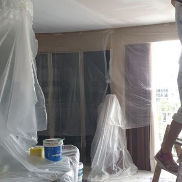 Habitación preparada para pintar; lonas de plástico cubren muebles y ventanas. Alguien sube a una escalera.