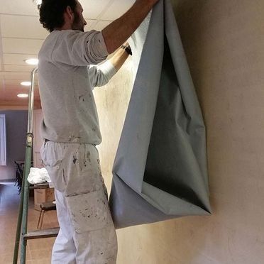 Hombre colgando una gran sábana gris en una pared de color claro, de pie sobre una escalera de mano.