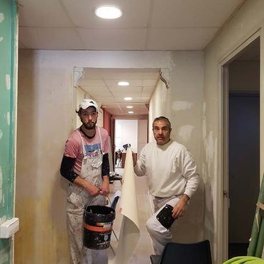 Dos pintores en un pasillo, uno sosteniendo un bote de pintura, el otro con papel tapiz.