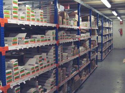 Estanterías de un almacén repletas de cajas y productos embalados. Estanterías azules y naranjas alinean las paredes, llenas de inventario.