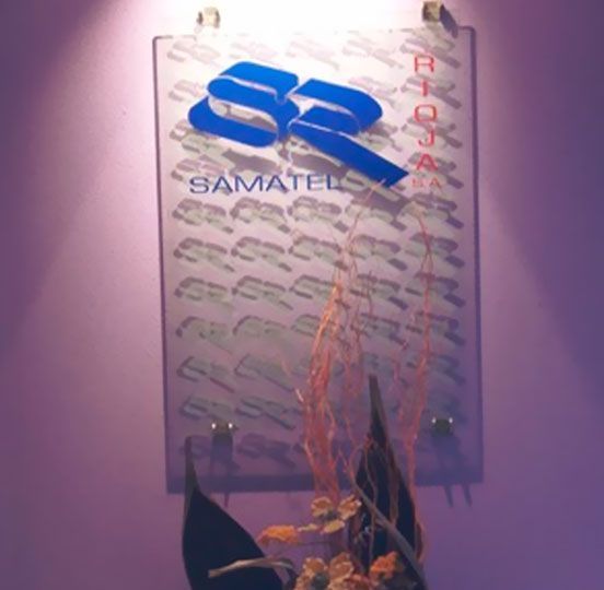 Rótulo para Samatel Rioja, logotipo azul sobre fondo transparente, montado sobre pared rosa con arreglo floral.