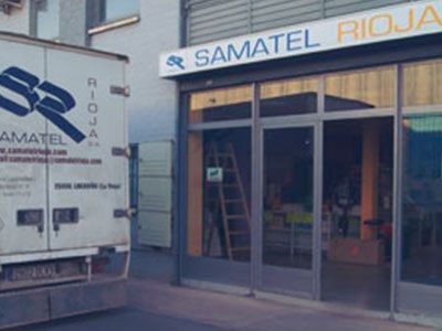 Un camión de reparto estacionado afuera de una tienda SAMATEL RIOJA con puertas de vidrio y una escalera visible en el interior.