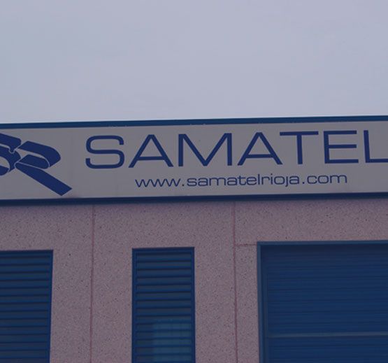 Letrero de SAMATEL, una empresa, con dirección de sitio web; montado sobre la entrada de un edificio, bajo un cielo nublado.