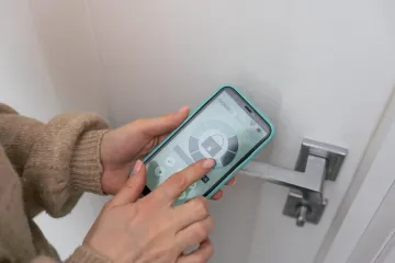 Manos utilizando un teléfono inteligente para controlar una cerradura inteligente en una puerta blanca.