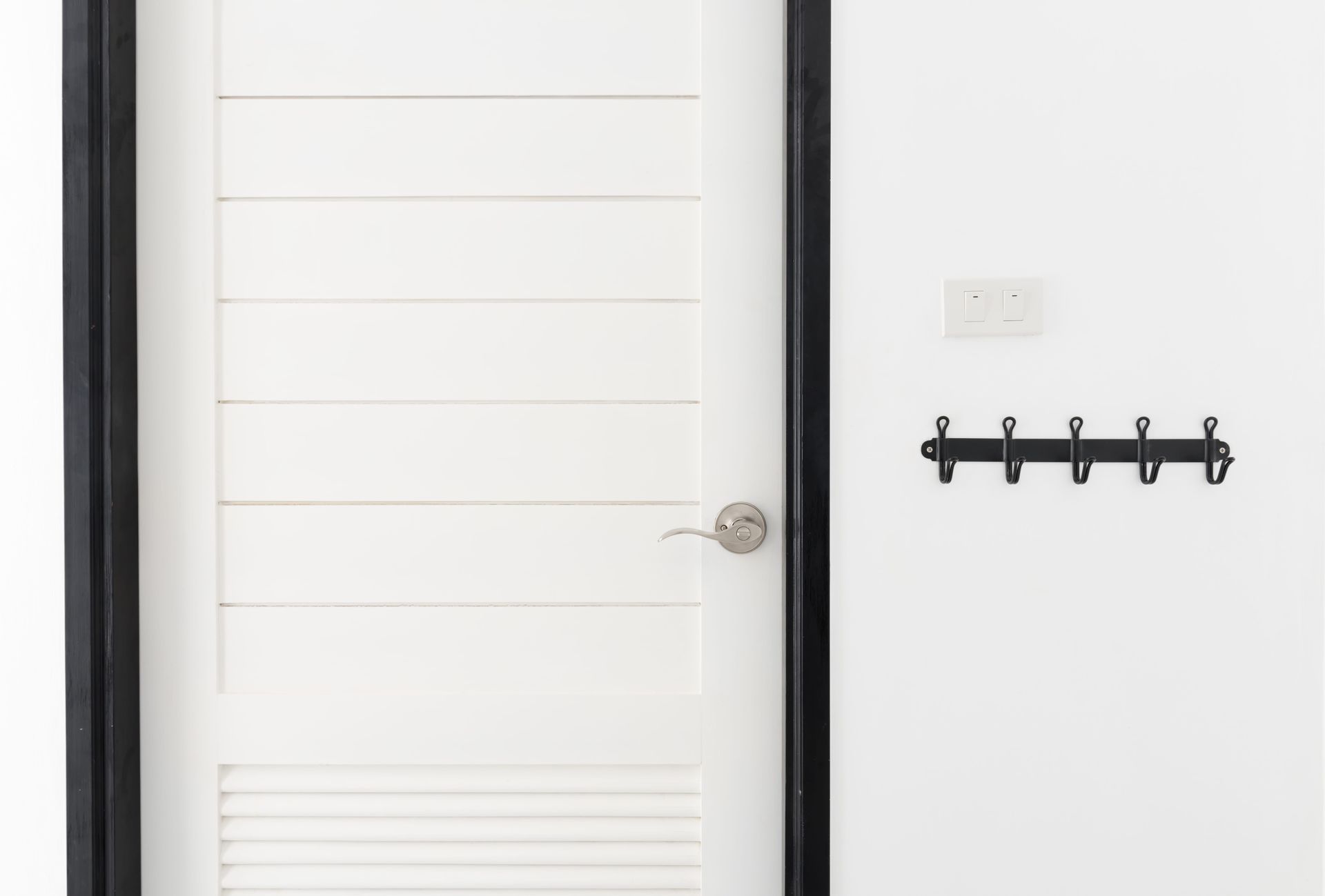 Puerta blanca con tirador plateado junto a un perchero negro montado en la pared y un interruptor de luz.