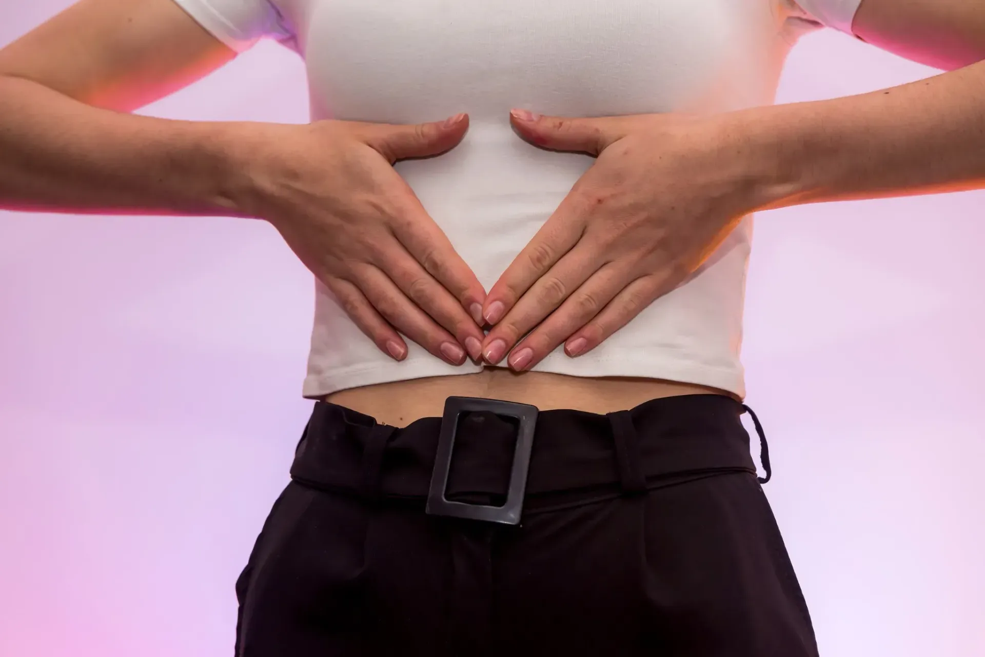 Las manos de una persona en forma de V sobre su estómago, sobre una camisa blanca y un pantalón negro.