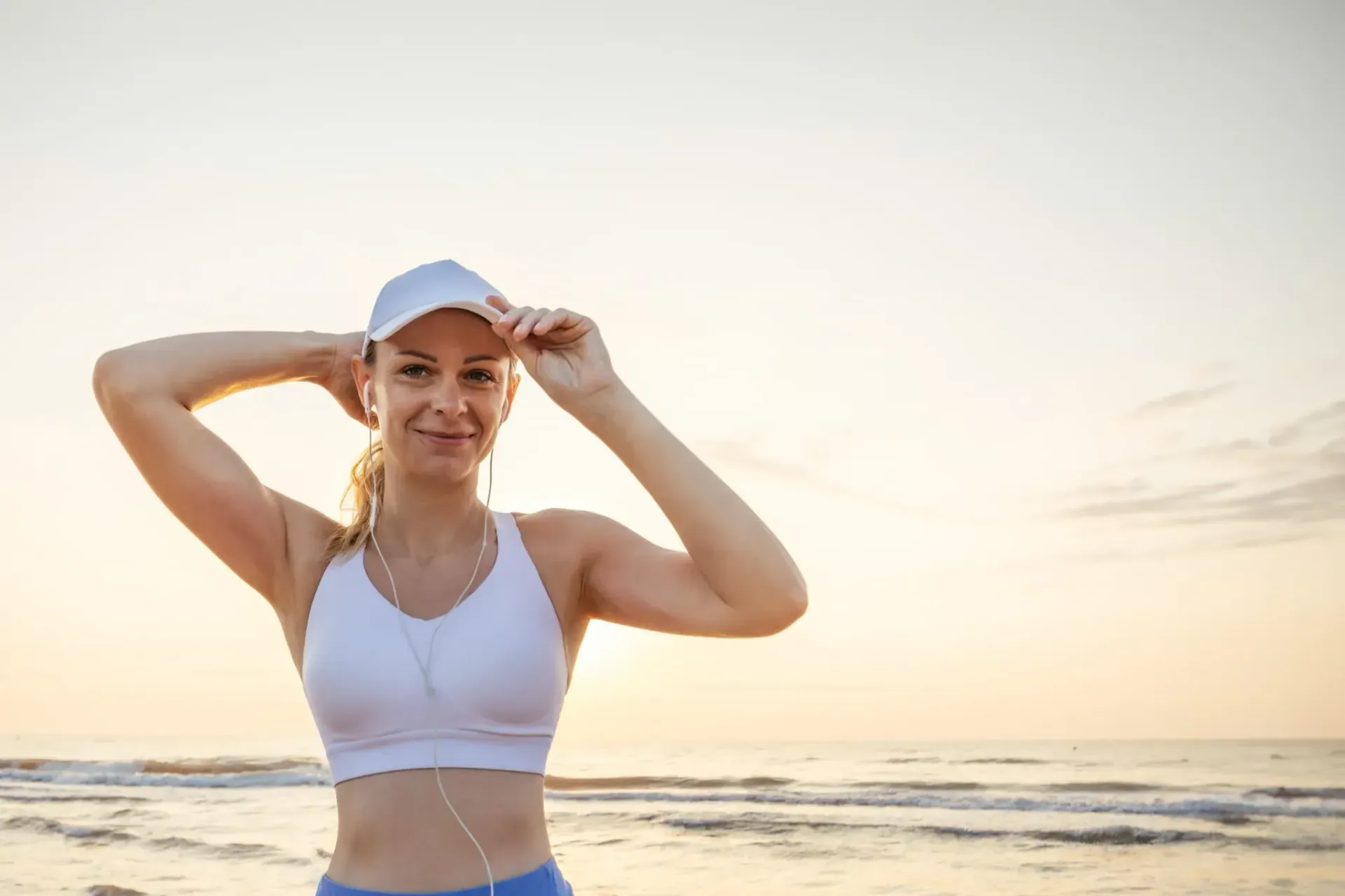 Mujer con ropa deportiva en la playa se ajusta la gorra; amanecer en el fondo.