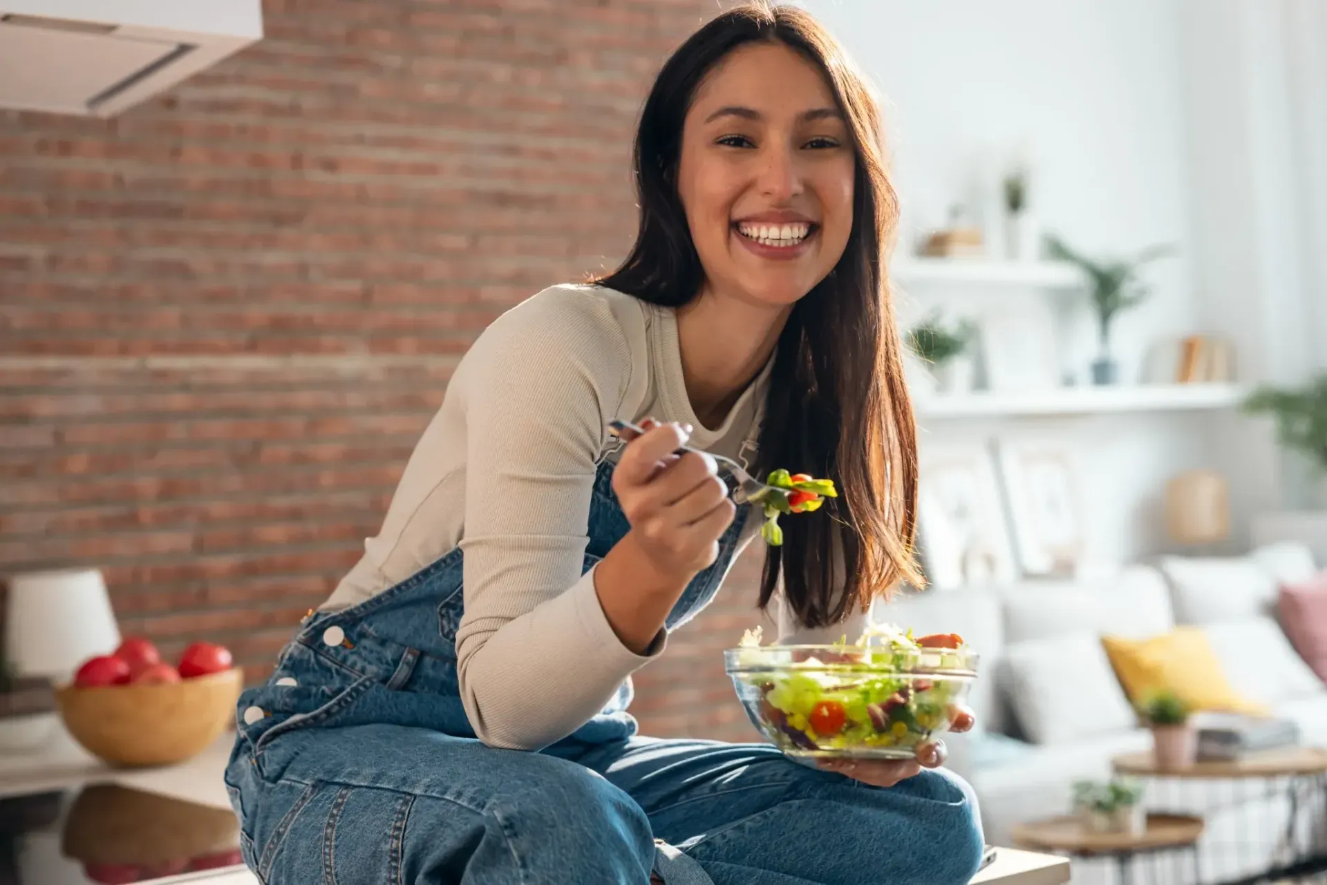 Mujer comiendo ensalada, sonriendo, en la cocina, vistiendo un mono de mezclilla.