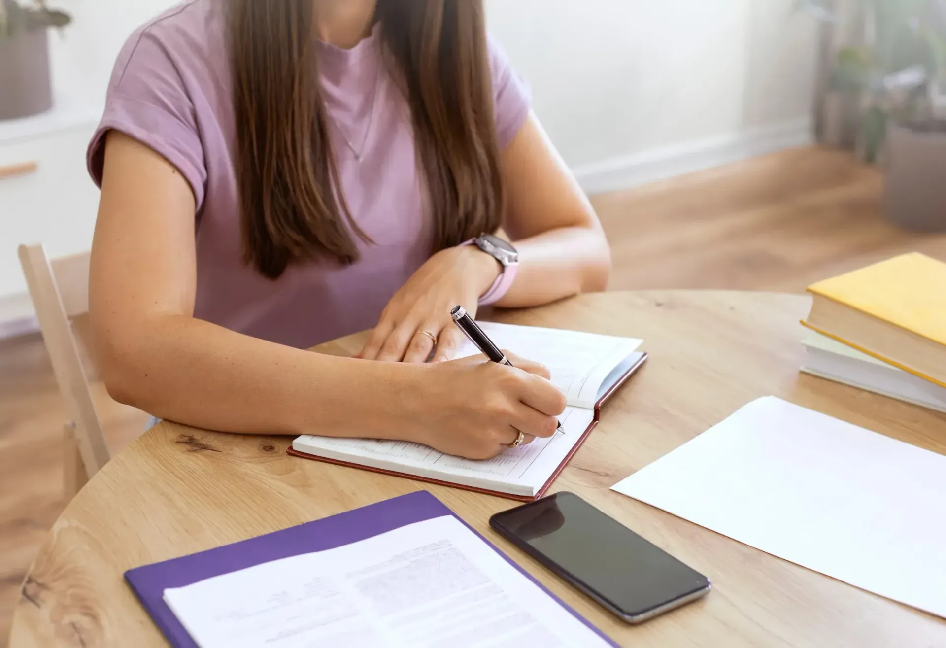 Mujer escribiendo en un cuaderno en una mesa de madera, con un teléfono y documentos cerca.